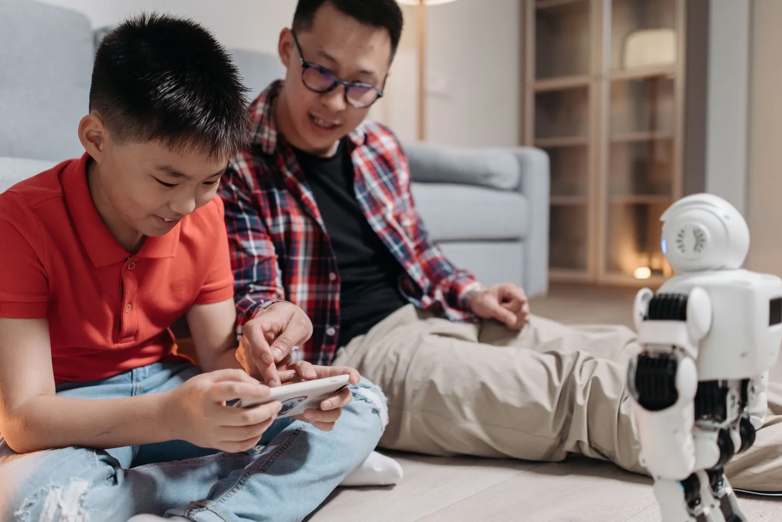 A father and his son sit on the floor, interacting playfully with a robot using a smartphone.